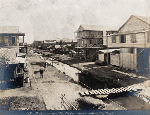 EU: Colón, Panama: a ditch runs down the middle of the unpaved street which is lined with wooden houses; a man on horseback is in the foreground. Photograph, (1907), Colón, Panama, 1907