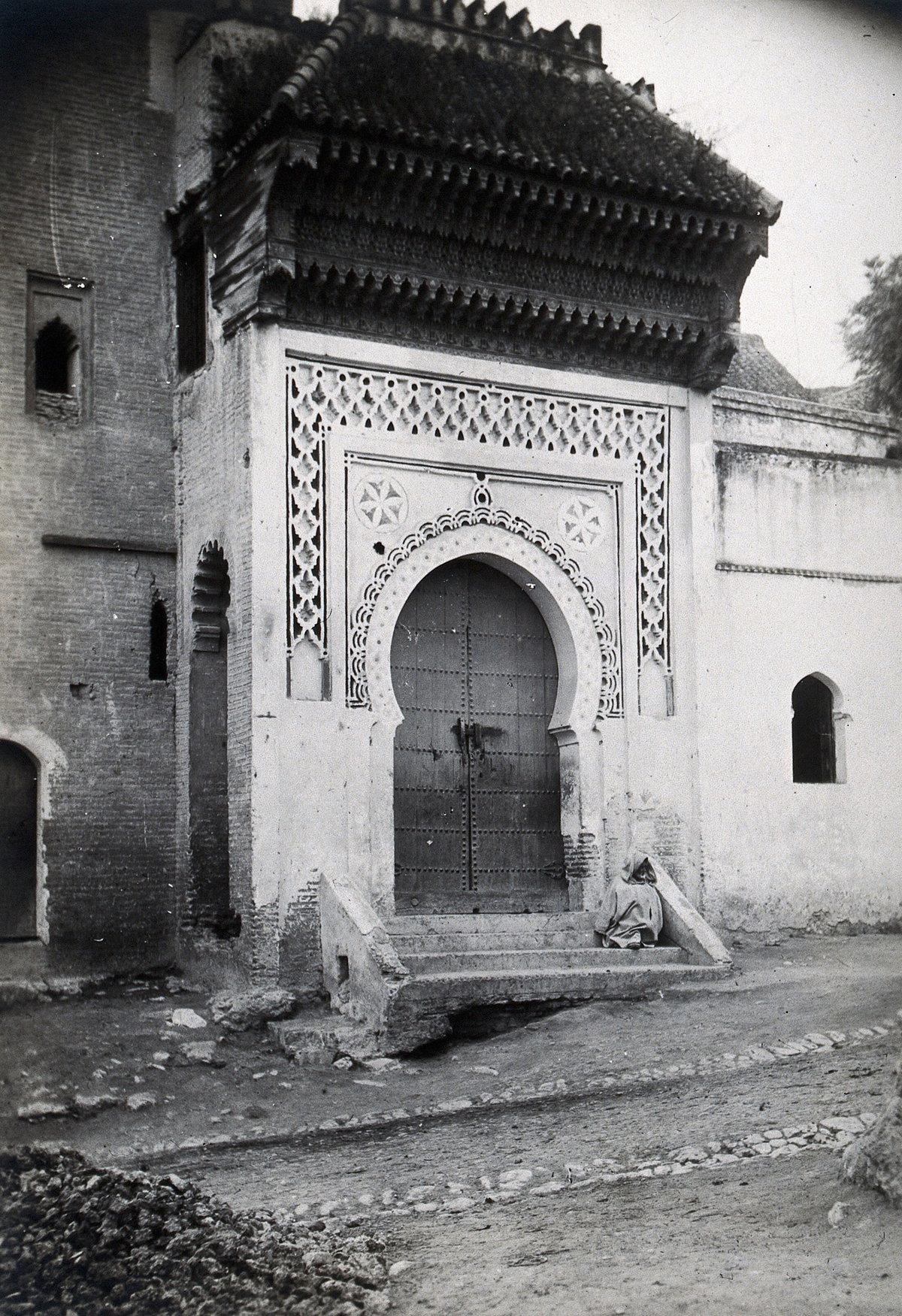 EU: Ornate doorway of the Medersa (Koranic college) of the Bab Ghissa Mosque, Fez, Morocco; male cloaked figure sitting on the steps. Photograph, ca. (1922), Fes, Morocco, 1922