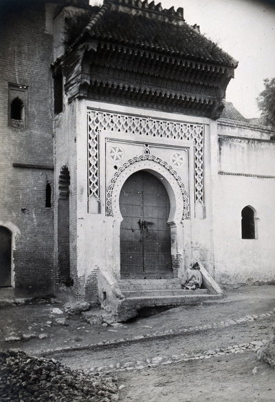 EU: Ornate doorway of the Medersa (Koranic college) of the Bab Ghissa Mosque, Fez, Morocco; male cloaked figure sitting on the steps. Photograph, ca. (1922), Fes, 1922