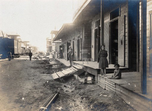EU: Colón, Panama: unpaved street lined with wooden houses; two children watch from a porch beside the open sewer. Photograph, (1906), Colón, Panama, 1906