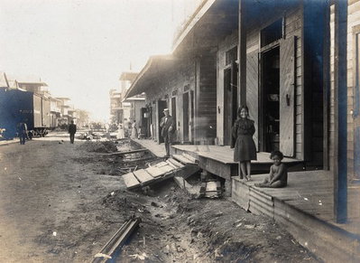 EU: Colón, Panama: unpaved street lined with wooden houses; two children watch from a porch beside the open sewer. Photograph, (1906), Colón, 1906