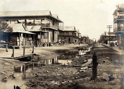 EU: Colón, Panama, before the sanitary works that were implemented during the construction of the Panama Canal: a ditch runs down the centre of a street lined with wooden houses. Photograph, Colón, 1908